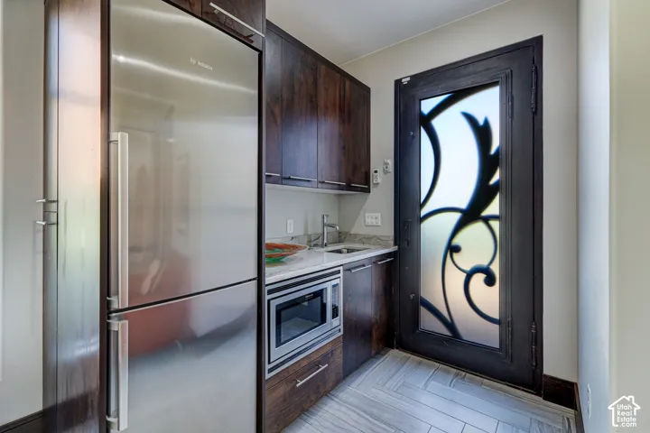 Stainless steel refrigerator and microwave, wood-finish cabinetry, stone countertop, decorative frosted glass door, and herringbone-patterned tile flooring