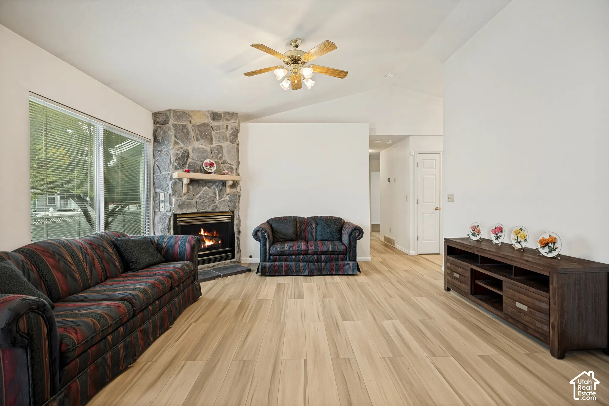 Vaulted ceiling, stone-clad fireplace, wood-finish flooring, ceiling fan, and large windows