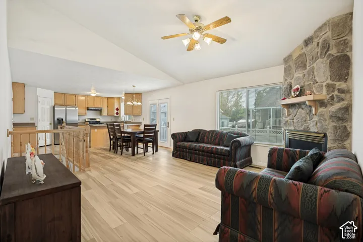 Vaulted ceiling, wood-finish flooring, stone fireplace, kitchen with light wood cabinetry, and stainless steel refrigerator