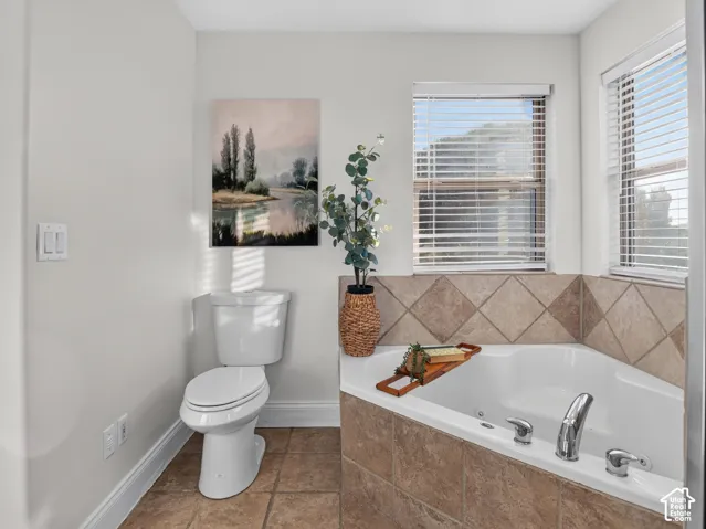 Corner soaking tub with tiled surround, dual windows with blinds, and neutral wall paint