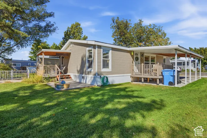 Beige horizontal siding exterior, covered porch with wooden railing, additional covered carport, concrete patio, and well-maintained lawn