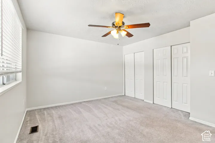 Neutral gray wall finishes, light gray carpeting, ceiling fan with light fixture, window with horizontal blinds, and paneled bi-fold closet doors