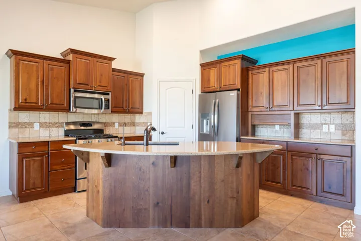 Kitchen featuring wood cabinetry, stainless steel appliances, a tiled backsplash, a central island with a sink, and tiled flooring