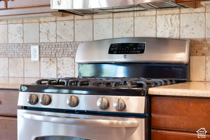 Stainless steel range with gas cooktop, stone tile backsplash with decorative accent, polished stone countertop, and wood cabinetry
