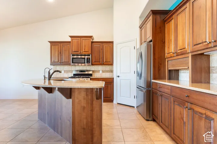 Kitchen with vaulted ceiling, wood-finish cabinetry, stainless steel appliances, tile flooring, and a curved island with an undermount sink