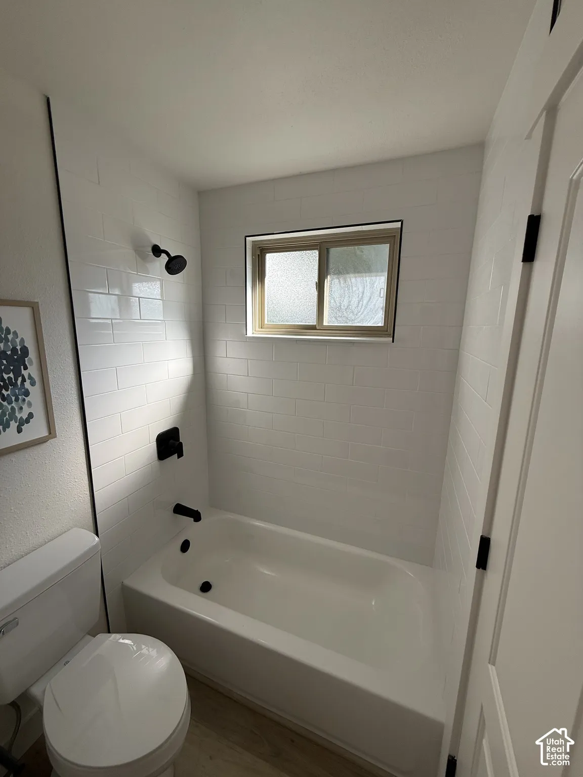 White subway tile shower surround with black fixtures, single glazed window, and wood-finish flooring