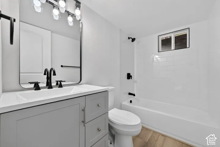 Modern bathroom featuring a gray vanity with white quartz countertop, matte black hardware and faucet, large framed mirror with integrated lighting, ceramic subway tile surround, soaking tub with glass enclosure, and wood-finish flooring
