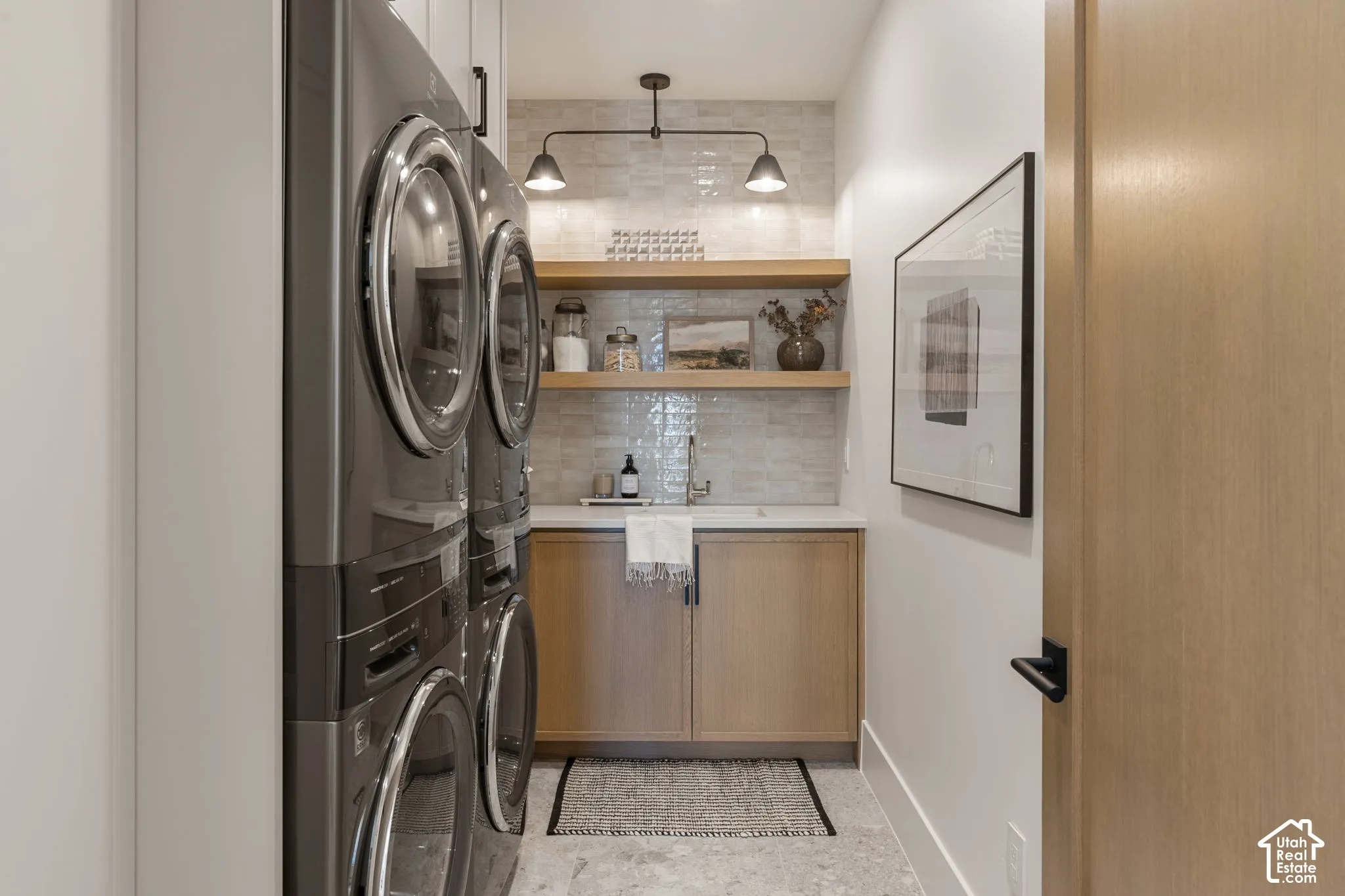 Stacked laundry appliances, light wood-finish cabinetry, floating wood-finish shelving, white countertop, and light-toned tile backsplash