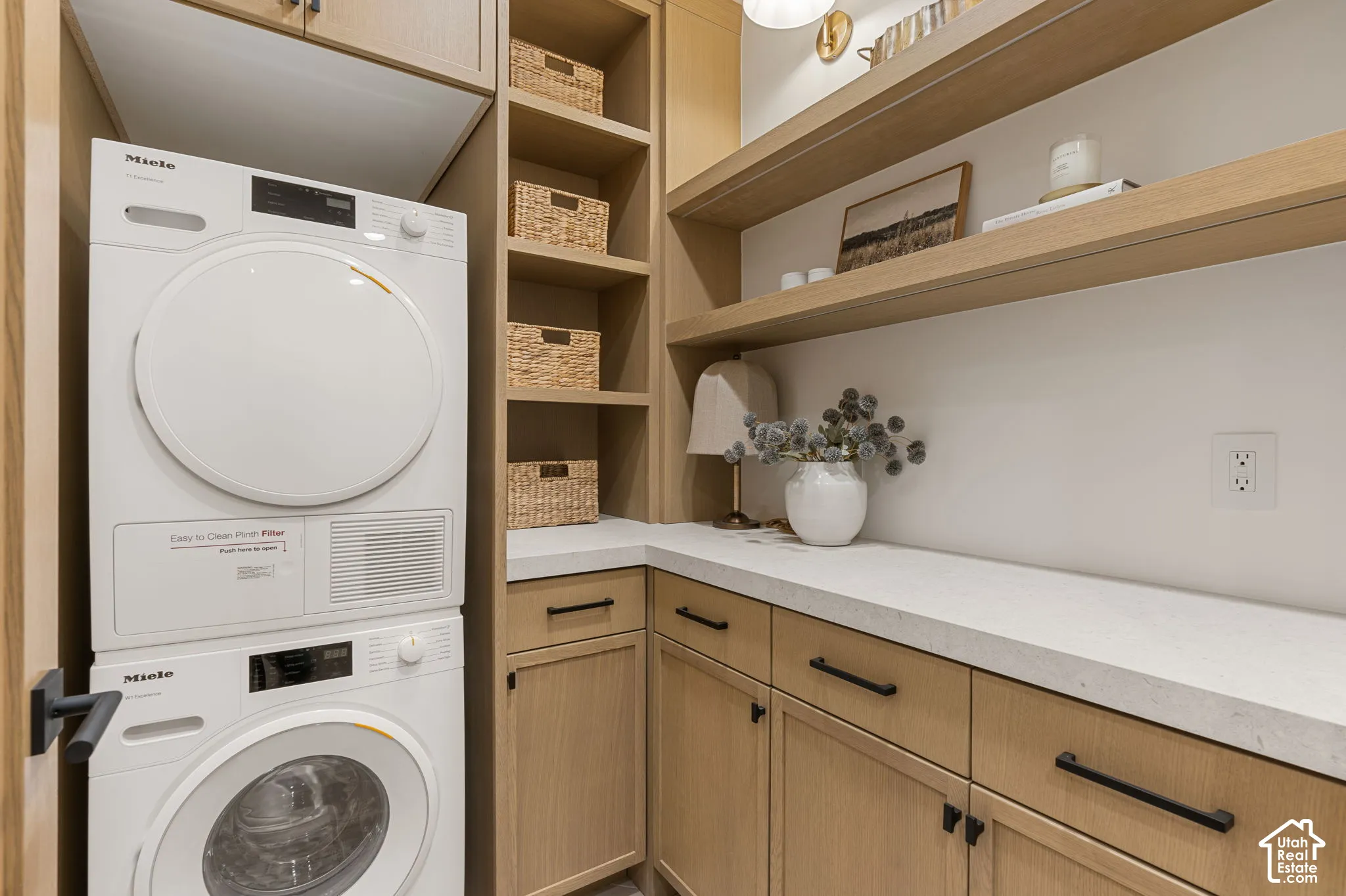 Stacked washer and dryer, light wood-finish cabinetry, solid surface countertop, open shelving, and built-in shelving units