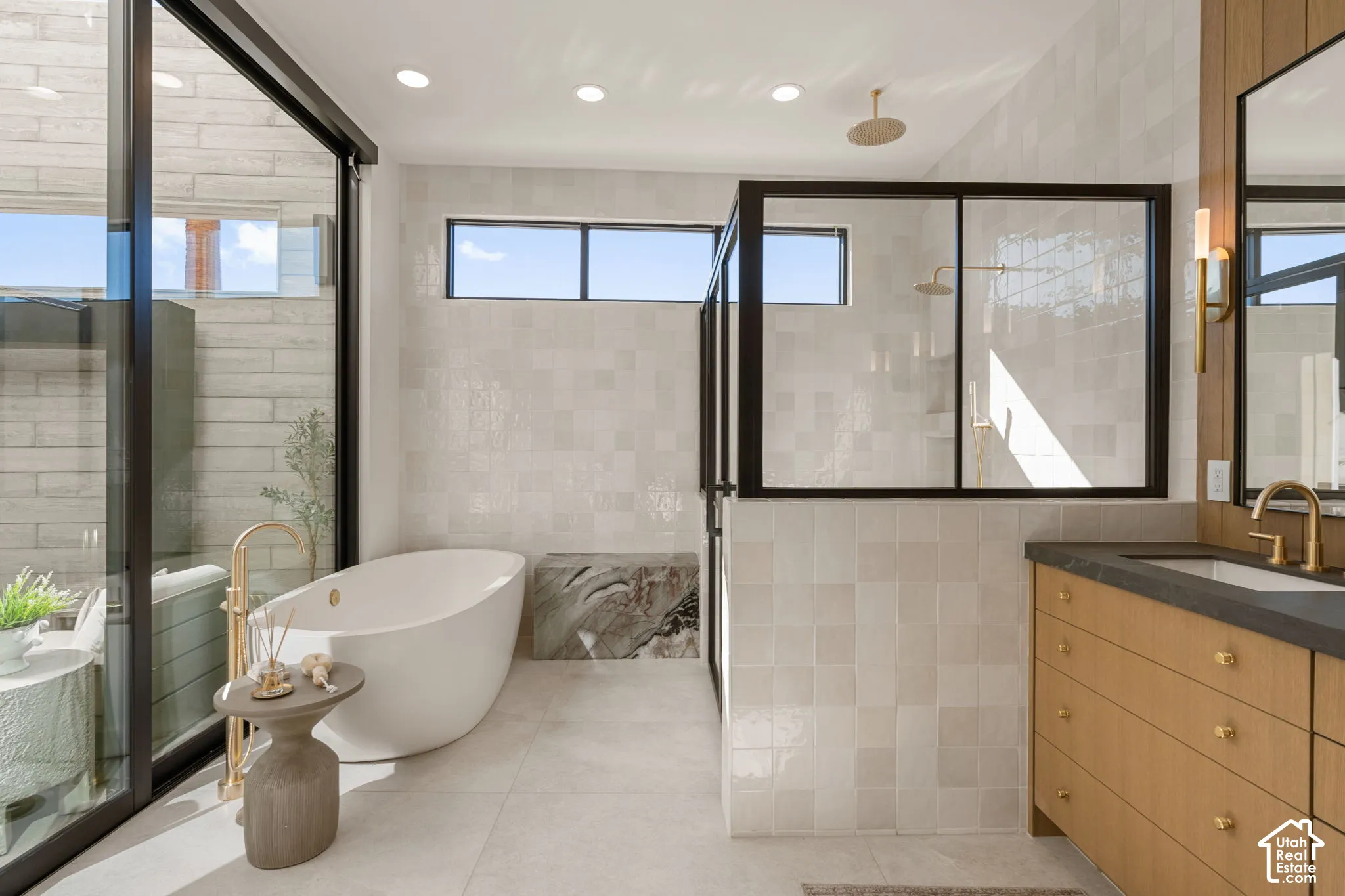 Freestanding soaking tub with gold-finish floor-mounted faucet, walk-in shower with black-framed glass enclosure, wood-finish vanity with dark countertop and undermount sink, and recessed lighting