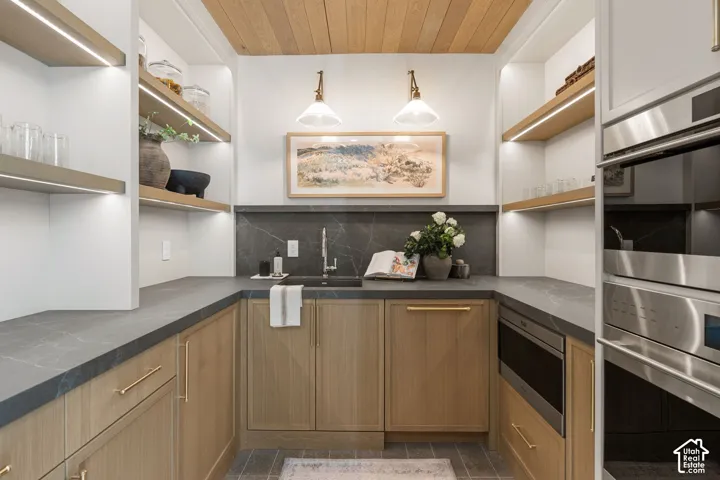 Integrated stainless steel double ovens, built-in microwave, wood-finish cabinetry, dark stone countertops with matching backsplash, and wood-paneled ceiling