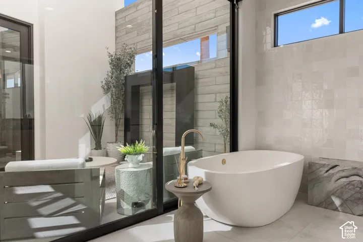 Freestanding soaking tub with gold-finish fixture, floor-to-ceiling glass sliding door, high transom window, and patterned tile wall