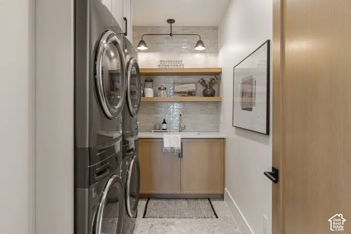 Stacked laundry appliances, light wood-finish cabinetry, floating wood-finish shelving, white countertop, and light-toned tile backsplash