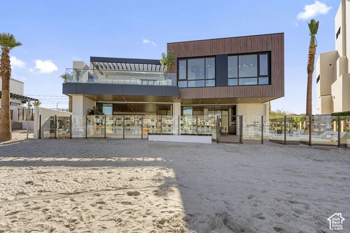 Modern architectural style with a flat roofline, wood-finish exterior accents, expansive windows, a second-story balcony with glass railings, and a ground-level patio area