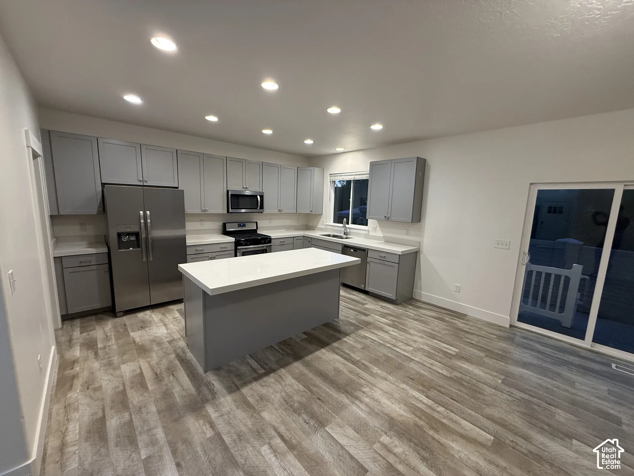 Modern kitchen featuring stainless steel appliances, grey cabinetry, a center island with a light-toned countertop, wood-finish flooring, and a sliding glass door
