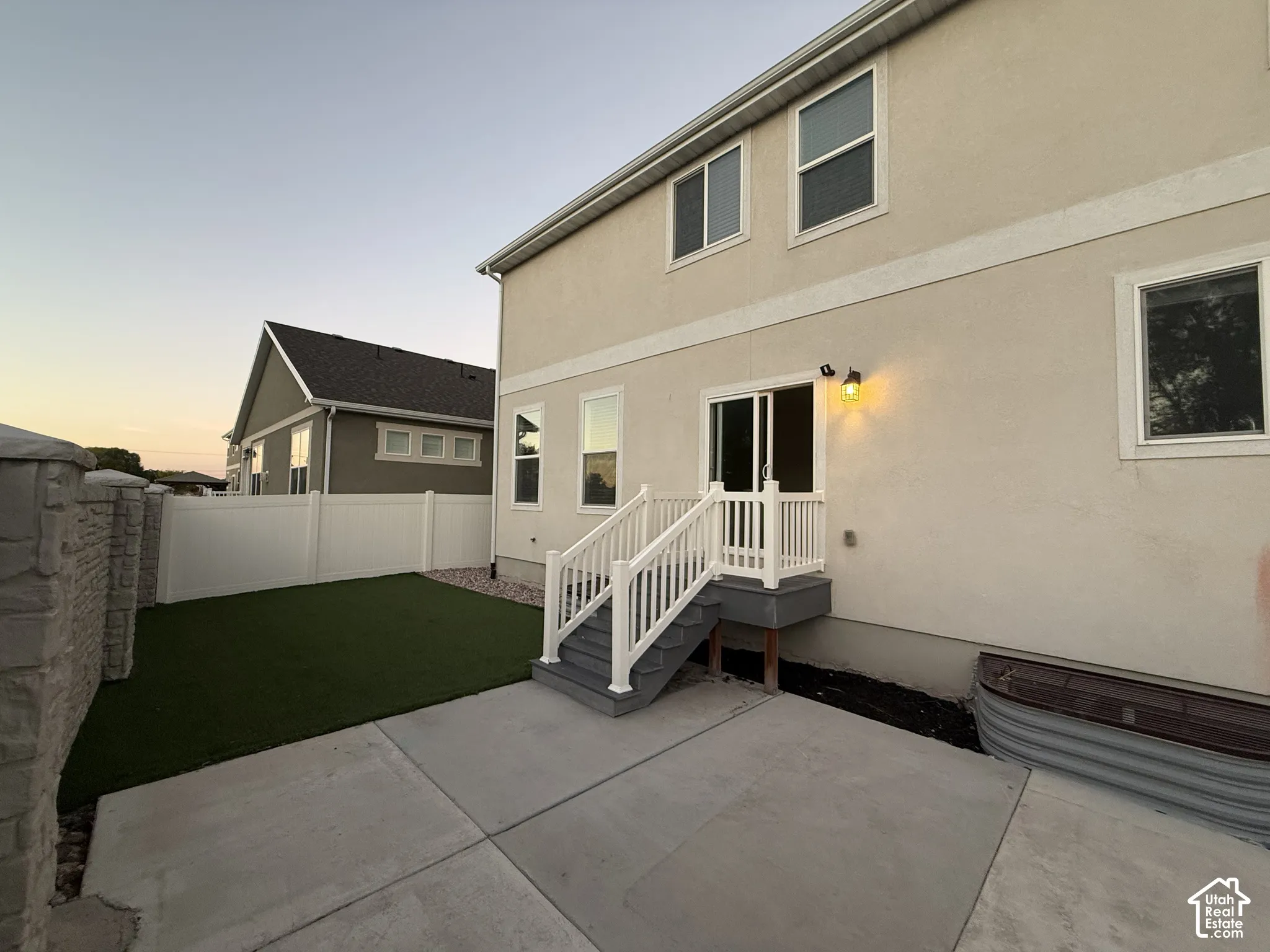 Rear exterior featuring a concrete patio, artificial turf, white privacy fencing, and a lower-level deck with white railings