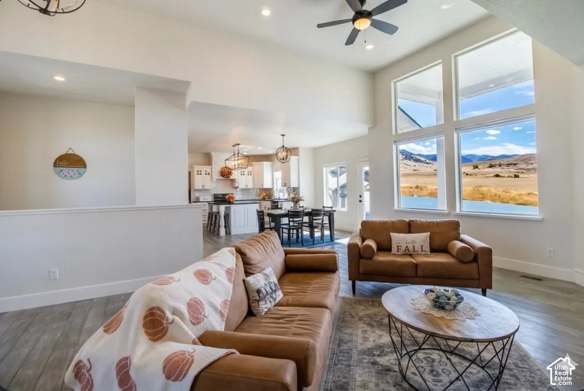 Open-concept living space featuring high ceilings with recessed lighting and ceiling fan, expansive windows framing scenic landscape vistas, wide-plank wood-finish flooring throughout, brown leather sectional seating, and a marble-topped oval coffee table