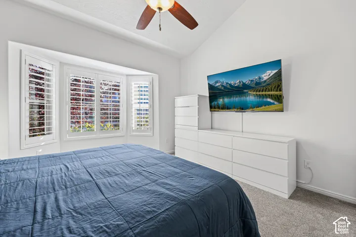 Vaulted ceiling with a ceiling fan, bay window featuring white plantation shutters, wall-mounted television, and neutral carpeting
