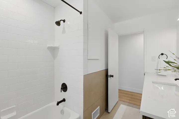 Subway tile shower surround with dark bronze fixtures, white bathtub, vanity with integrated sink, and wood-finish flooring