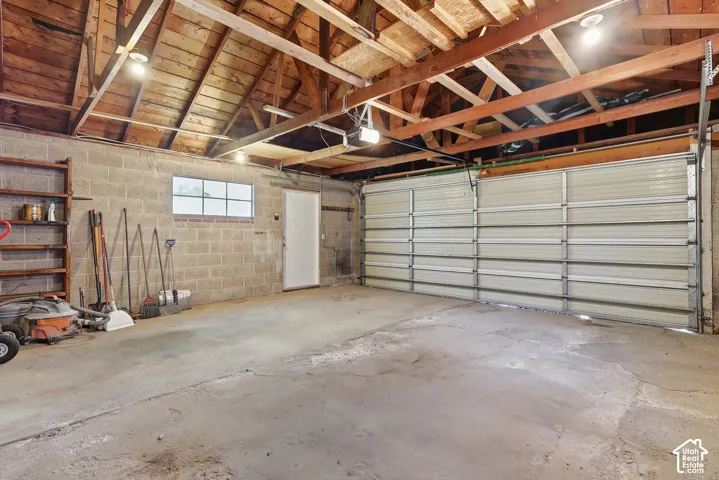 Concrete block walls, exposed wood beam ceiling, concrete flooring, overhead lighting, and an overhead garage door