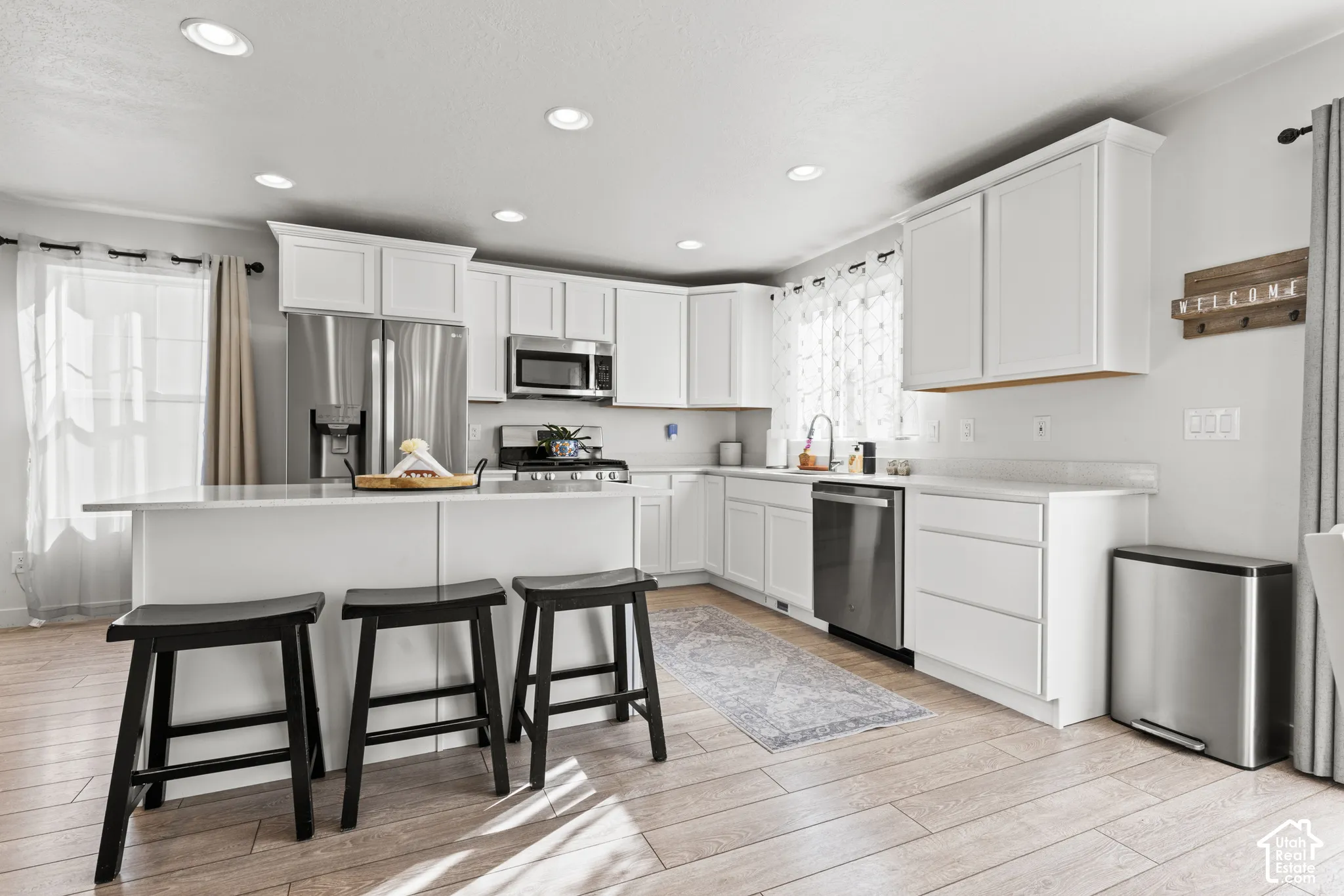 Recessed lighting illuminates a kitchen featuring wood-finish flooring, white cabinetry, a kitchen island with a white countertop, and stainless steel appliances
