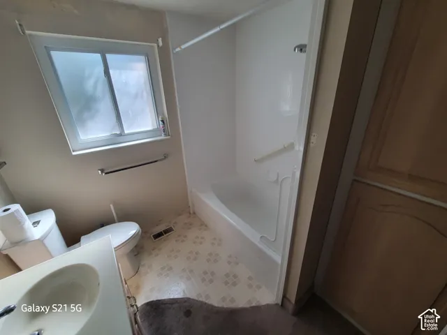 Bathroom featuring a single-pane window, white vanity with integrated sink, white toilet, and a combination shower/tub with grab bars