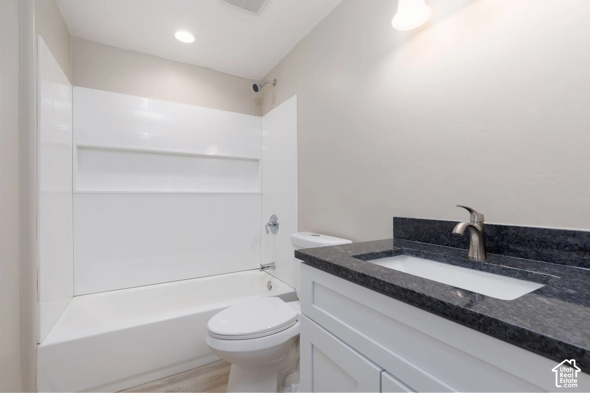 White bathtub with an integrated shower, white toilet, white vanity with a dark countertop, and a brushed nickel faucet