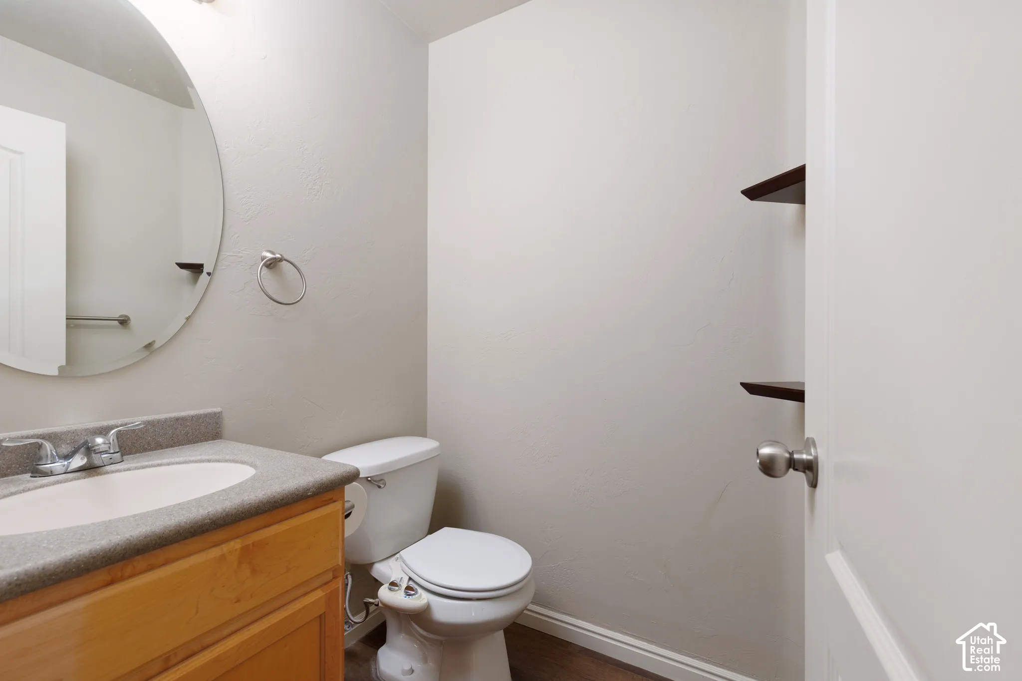 Wood-finish flooring, wood-finish vanity with integrated sink, chrome faucet, wall-mounted mirror, and floating shelving