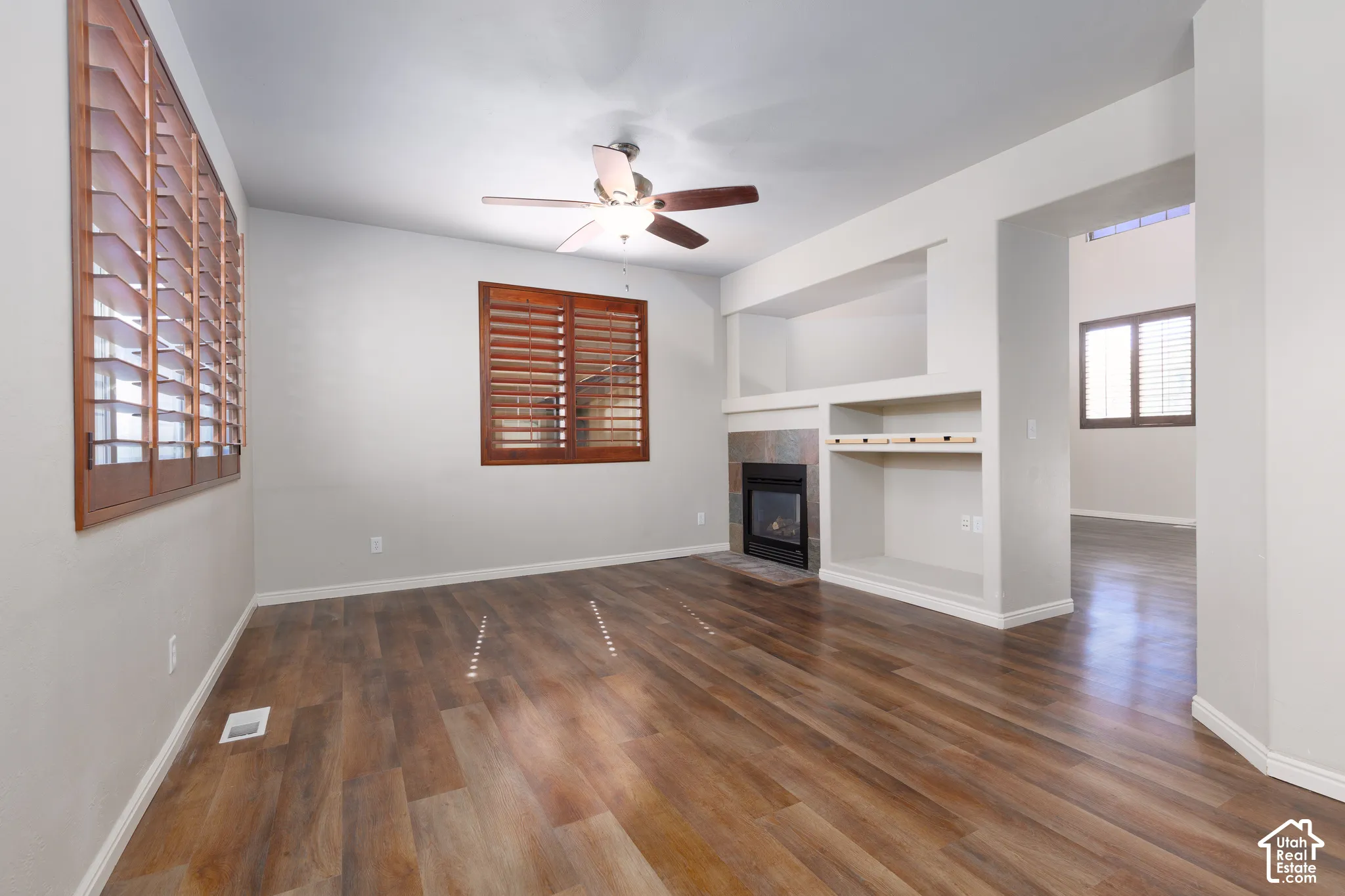 Wood-finish flooring, built-in shelving, fireplace with stone surround, ceiling fan, and wood plantation shutters