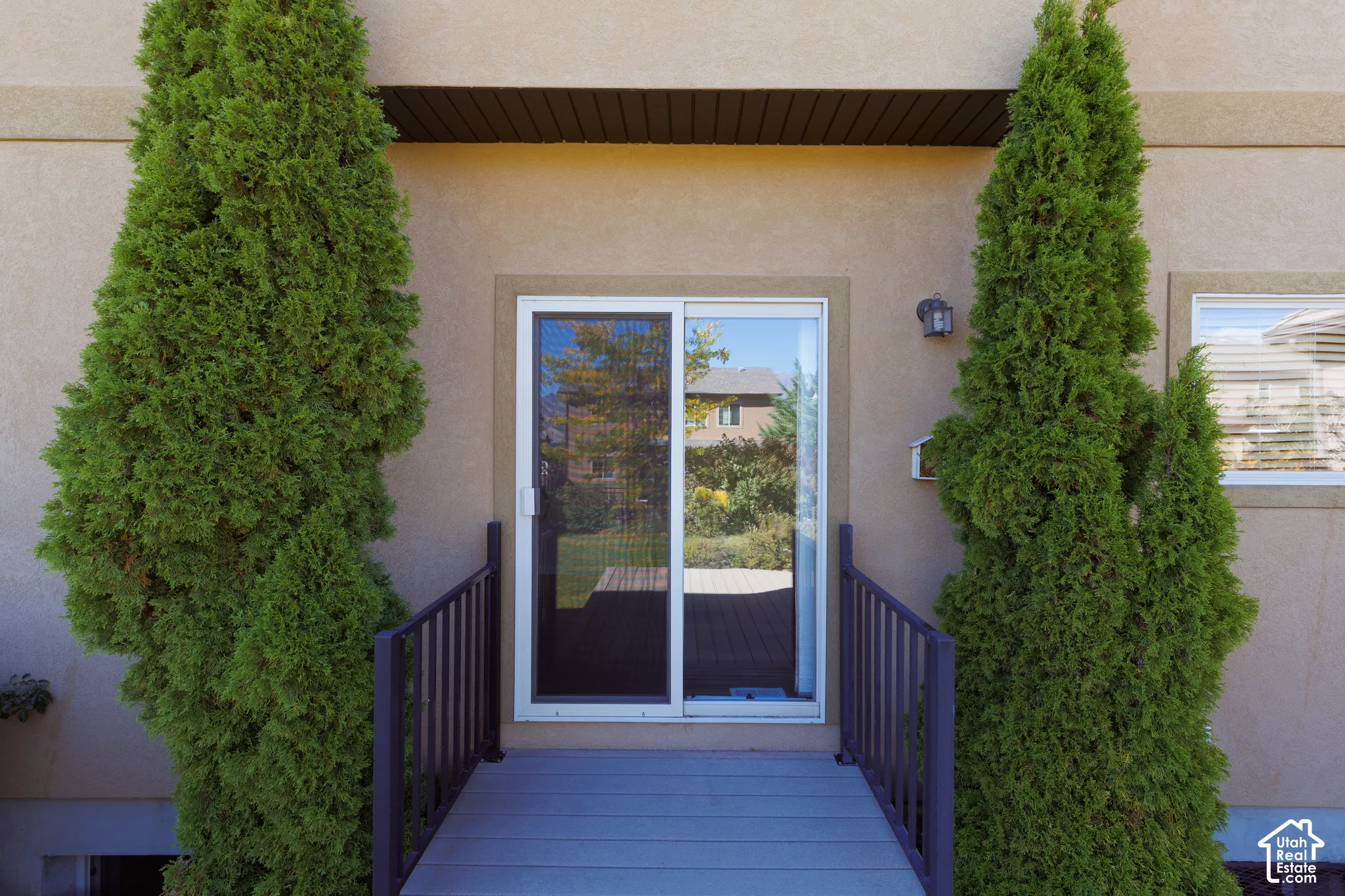 Sliding glass door, built-in outdoor lighting fixture, stucco exterior finish, window with blinds, and dark railing