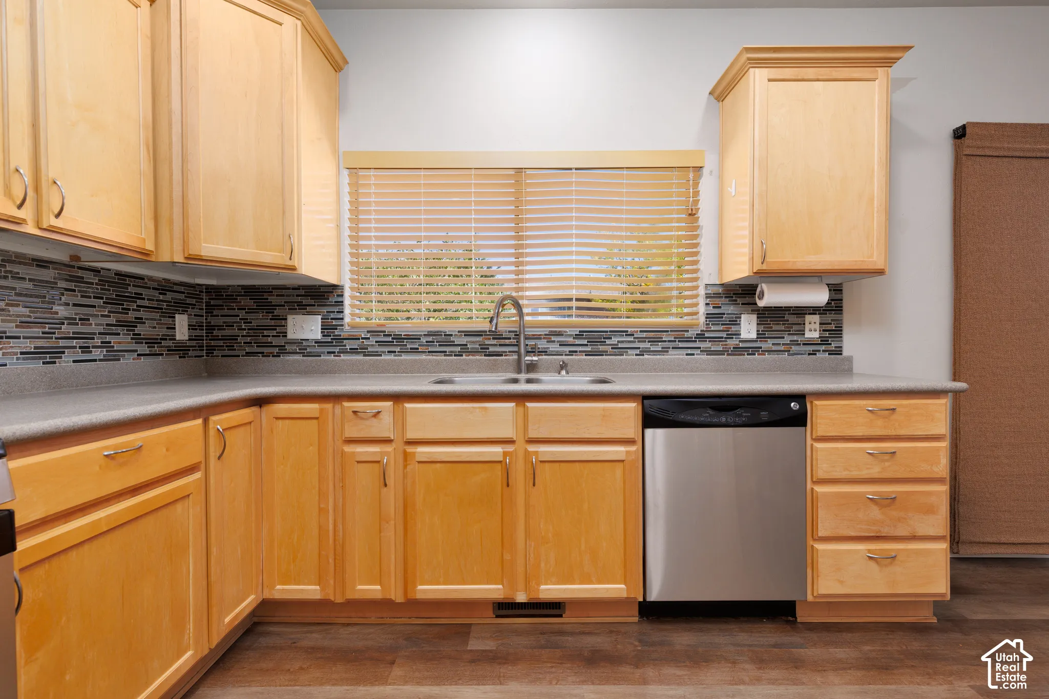 L-shaped countertop with a tile backsplash, wood-finish cabinetry, stainless steel dishwasher, and a window with blinds