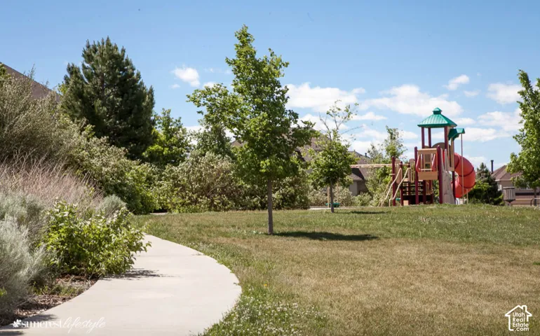 Community green space featuring a curved concrete pathway, a playground structure with a slide, mature trees, and landscaping