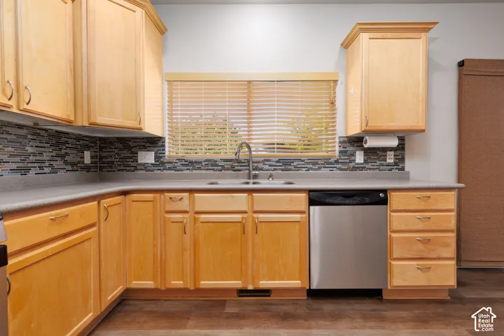 L-shaped countertop with a tile backsplash, wood-finish cabinetry, stainless steel dishwasher, and a window with blinds