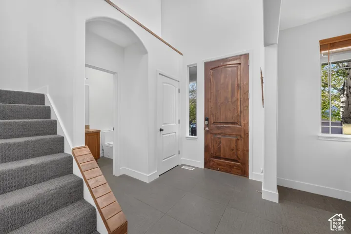 Hardwood entry door, gray tile flooring, carpeted staircase, and arched hallway