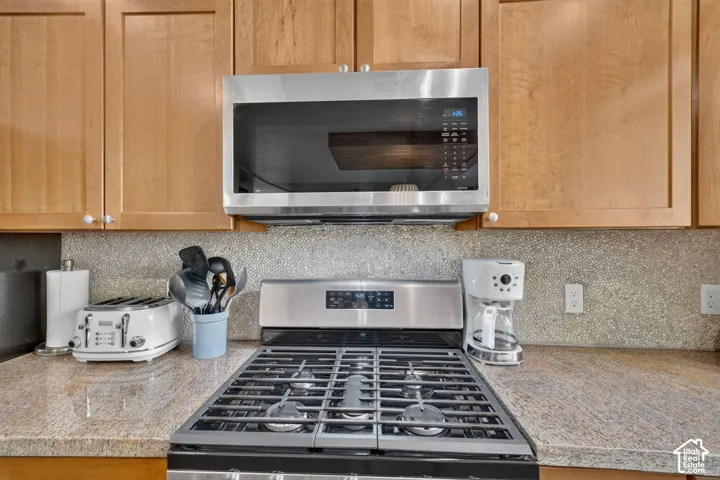 Stainless steel range with gas burners, built-in microwave, wood cabinetry, granite-style countertop, and textured backsplash