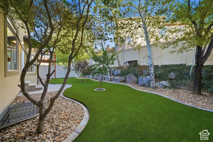 Landscaped backyard featuring an artificial turf lawn, mature trees, decorative rock beds, and a privacy fence