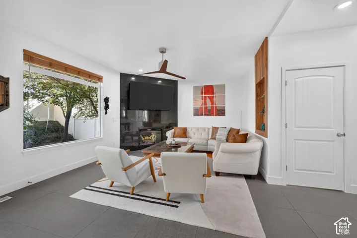 Spacious living area featuring a large window, dark tile flooring, a built-in fireplace with a dark surround, a ceiling fan, and a wood-paneled accent wall with integrated shelving
