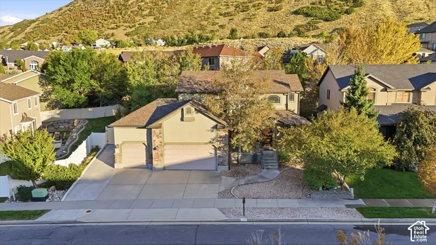 Two-car garage with stone accents, concrete driveway, front yard with mature trees, and a mountain backdrop