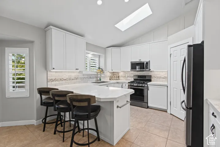 Vaulted ceiling with skylight, white shaker cabinetry, stainless steel appliances, white tile backsplash, and an island with seating