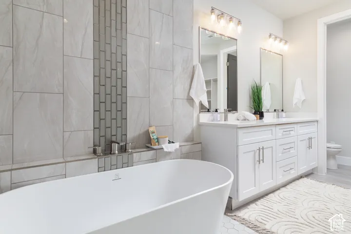 Freestanding soaking tub, floor-to-ceiling tile surround with accent mosaic, dual vanity with white cabinetry, integrated sinks, and individual mirrors