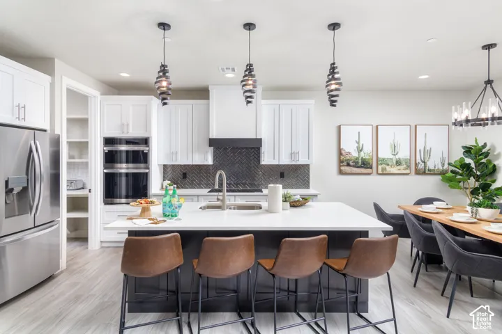 Modern kitchen featuring white cabinetry, stainless steel appliances, a center island with sink, herringbone tile backsplash, and wood-finish flooring