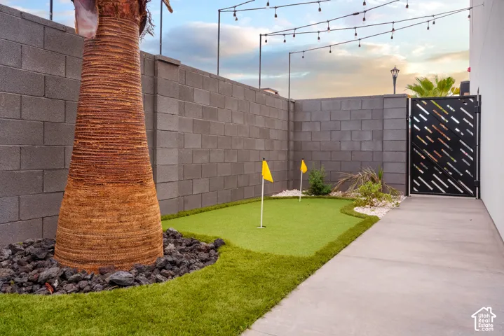 Paved side yard with a putting green, decorative metal gate, concrete block wall, and integrated string lighting