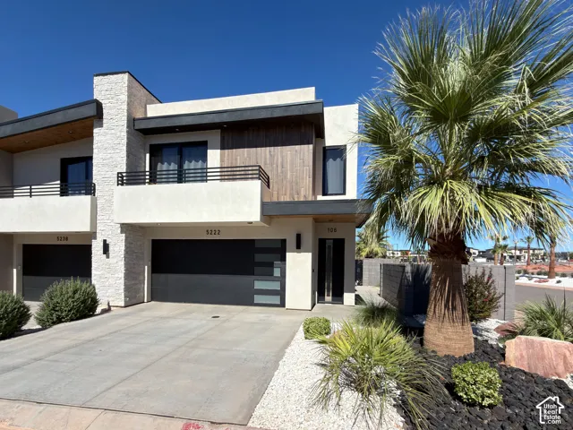 Modern exterior with stucco and stone veneer accents, wood-paneled upper facade, private balcony with black railings, contemporary garage door, and desert-adapted landscaping