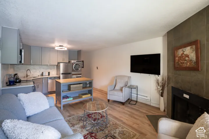Open-concept living space with wood-finish flooring, a stone-tiled fireplace, and a kitchen featuring grey cabinetry, stainless steel appliances, and a tiled backsplash