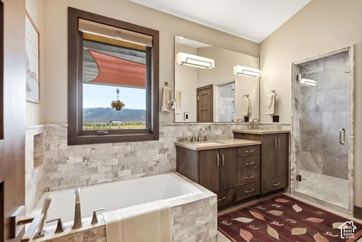 Built-in soaking tub, wood-finish window trim, stone-tiled walls, dual vanity with undermount sinks, and a glass-enclosed shower