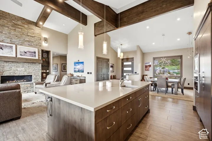 Vaulted ceilings with exposed wood beams, a stone-faced fireplace, a large kitchen island with a built-in sink, wood-finish flooring, and integrated kitchen appliances