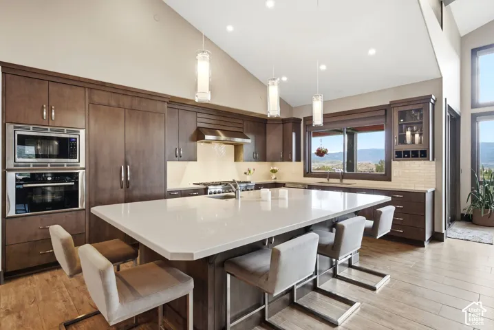 Vaulted ceiling with recessed lighting, large central island with light-toned countertop, wood-finish flooring, built-in double oven, and integrated cabinetry
