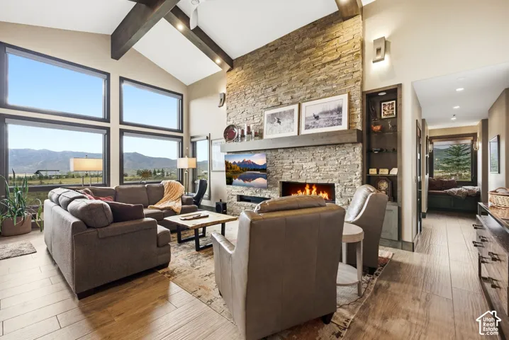 Vaulted ceiling with exposed wood beams, stone-clad fireplace, built-in shelving, wood-finish flooring, and large picture windows
