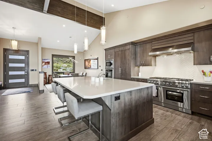 Open-concept kitchen featuring an expansive island with seating, integrated stainless steel appliances, a herringbone tile backsplash, and wood-finish flooring