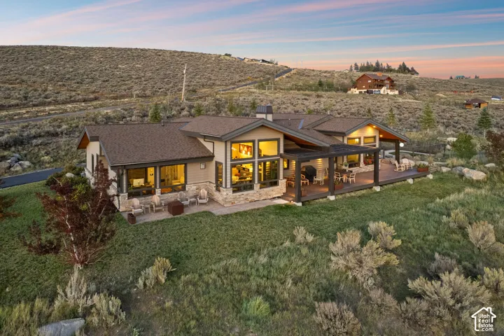 Spacious rear deck, covered patio area, large windows, stone facade accents, and a sloped, natural landscape